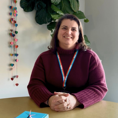 Teresa smiling seated at a table with a pile of books in front and a tall leafy green plant behind her.
