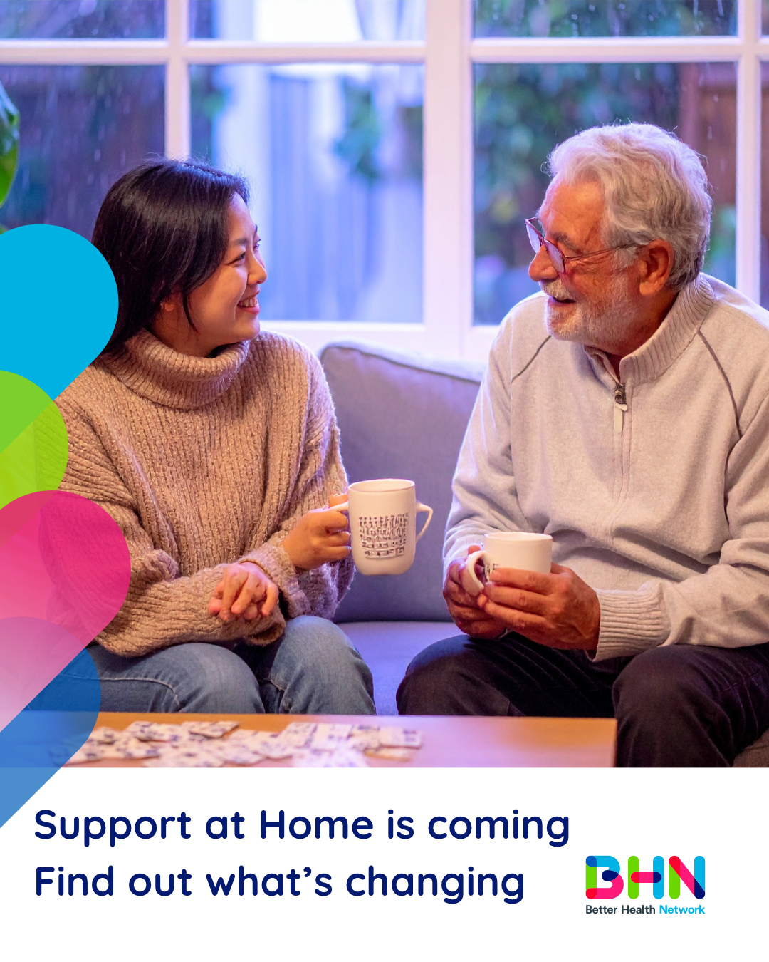 Support at Home is coming post Smiling woman and older man sitting on couch holding coffee mugs and talking. Puzzle pieces spread out on coffee table.